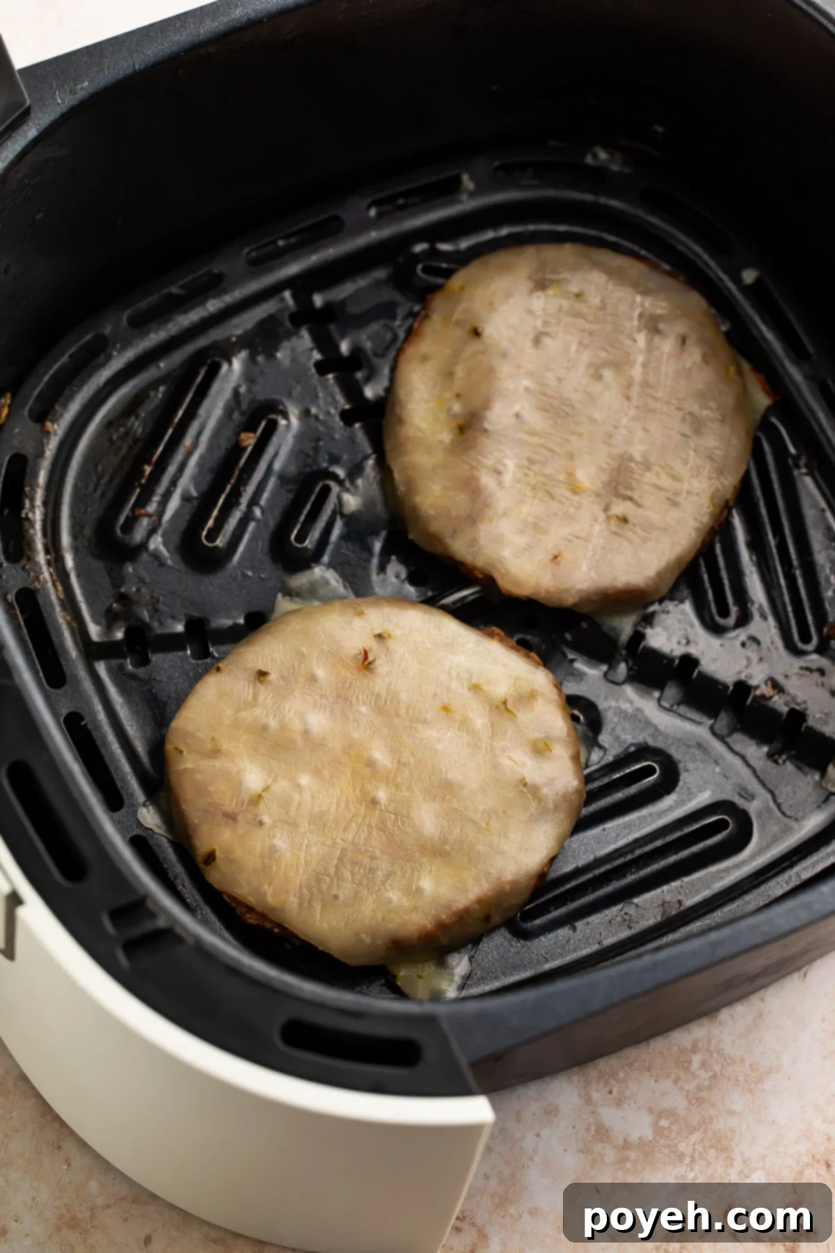 Two frozen burger patties topped with cheese cooking in an air fryer basket, showing the efficiency of the air frying method.