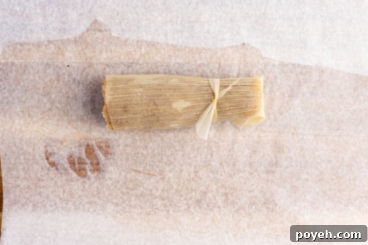 A perfectly rolled keto tamale, securely tied with a corn husk strip, resting on a parchment paper lined cutting board.