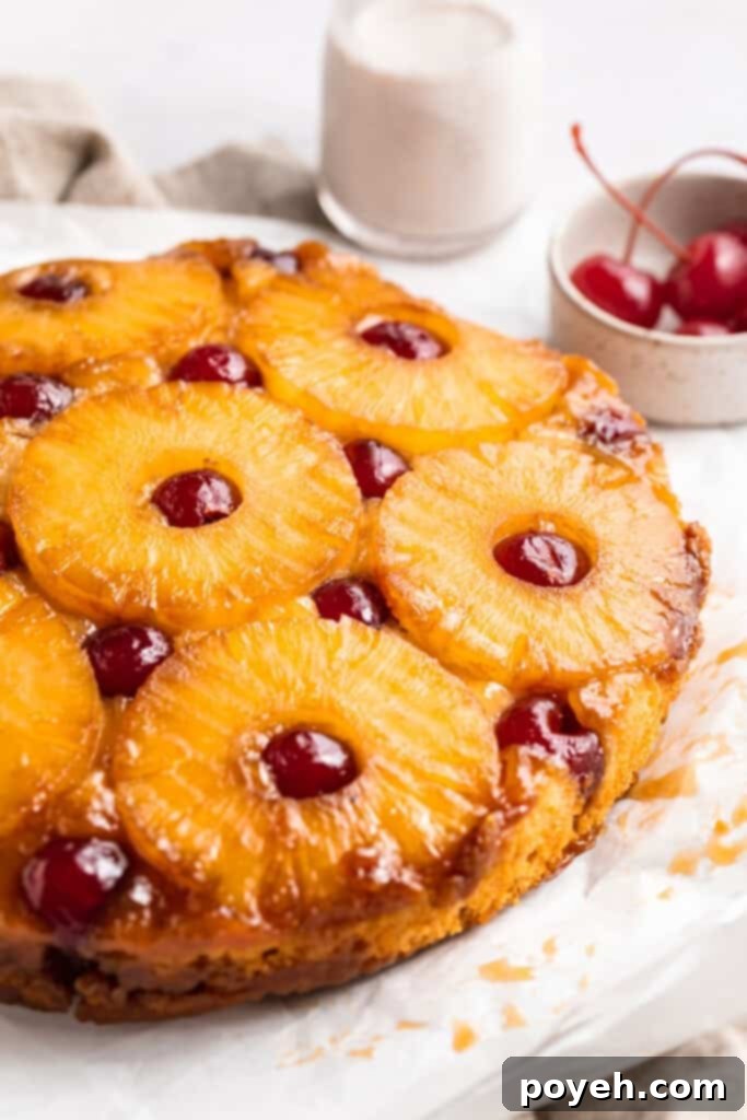 Close-up, 3/4 angle shot of a vibrant vegan pineapple upside down cake, adorned with glistening pineapple rings and bright red maraschino cherries, resting elegantly on a pristine white table.