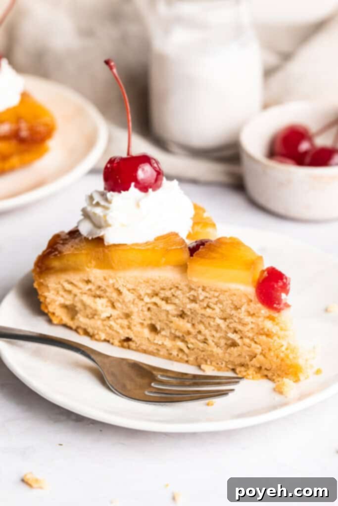 A perfectly portioned wedge-shaped slice of vegan pineapple upside down cake, adorned with a dollop of fluffy vegan whipped cream and a bright red maraschino cherry, presented on a small, elegant dessert plate.