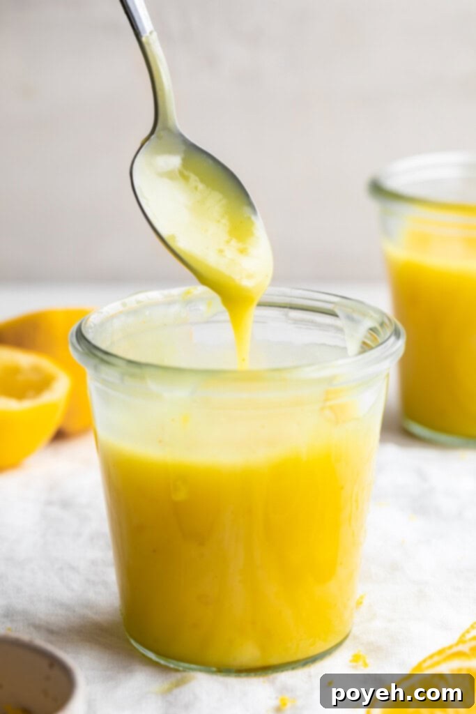A beautifully set glass jar filled with vibrant yellow vegan lemon curd, with a spoon gently pouring more curd into the jar, highlighting its smooth and creamy texture.