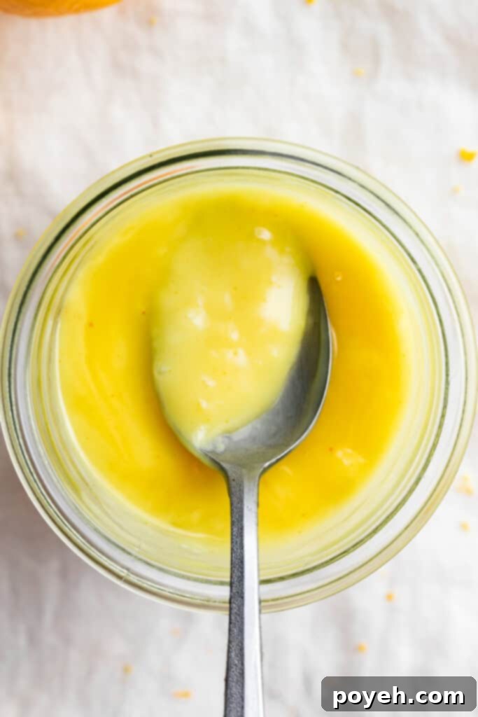An overhead shot showcasing a glass jar brimming with smooth, bright yellow vegan lemon curd, with a spoon resting elegantly beside it.