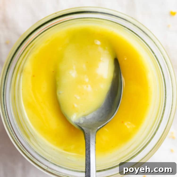 Overhead view of a jar of vibrant yellow vegan lemon curd with a spoon, set against a stylish background.