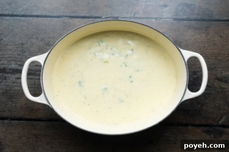Overhead view of a large soup pot containing sautéed vegetables, flour, and now heavy cream, being whisked into a smooth, creamy base.