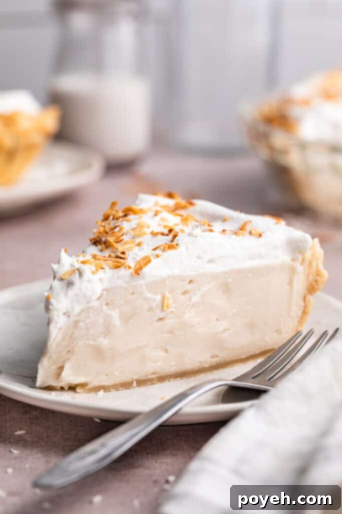 A wedge sliced of vegan coconut cream pie topped with coconut whipped cream and coconut shavings on a neutral plate with a fork, ready to be enjoyed.
