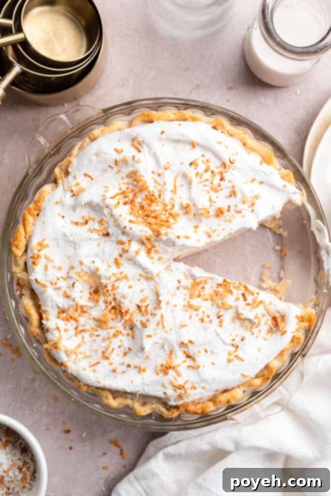 Overhead view of a whole vegan coconut cream pie topped with toasted coconut, with one slice missing from the plate, showcasing its perfection.