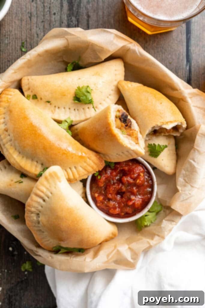 Overhead view of a beautiful array of baked vegan empanadas, perfectly golden, arranged in a rustic bowl next to a small ramekin of rich red salsa, ready for dipping.