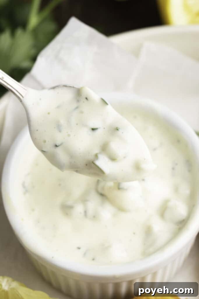 Vegan tartar sauce being lifted out of a small white ramekin on a silver spoon, showcasing its creamy texture and visible flecks of herbs and pickles.