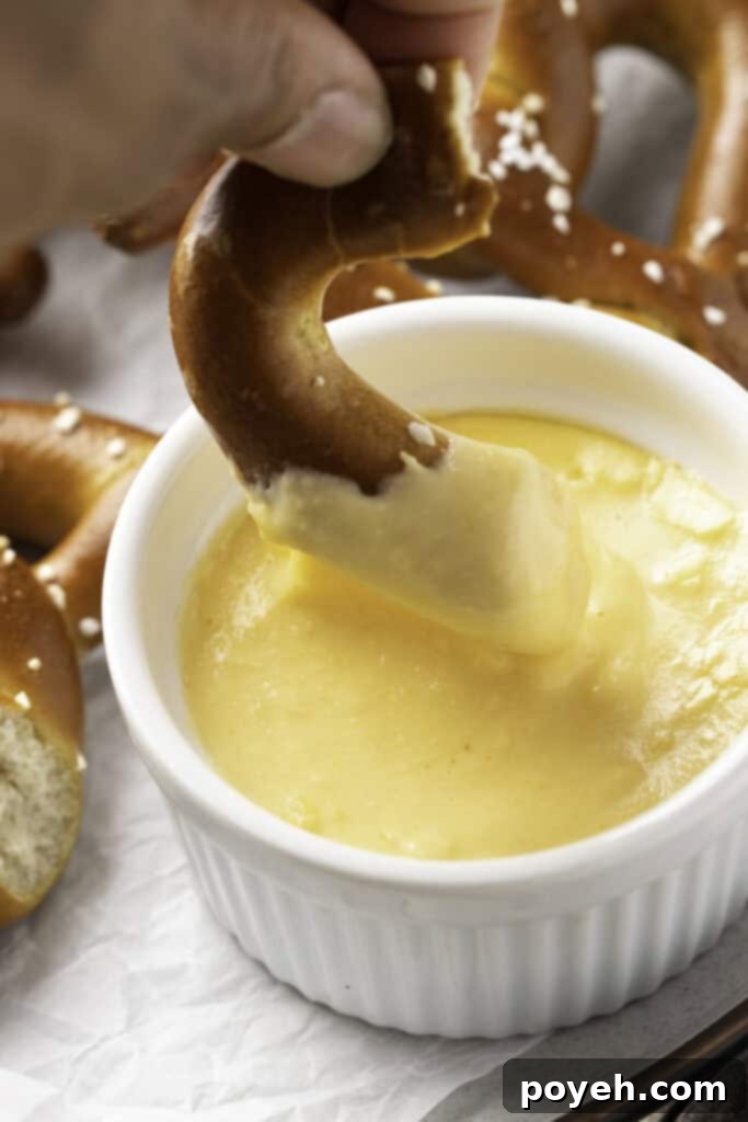 A close-up shot of a piece of salted, twisted, air fryer frozen pretzel being dipped into a small ramekin of creamy cheese sauce.