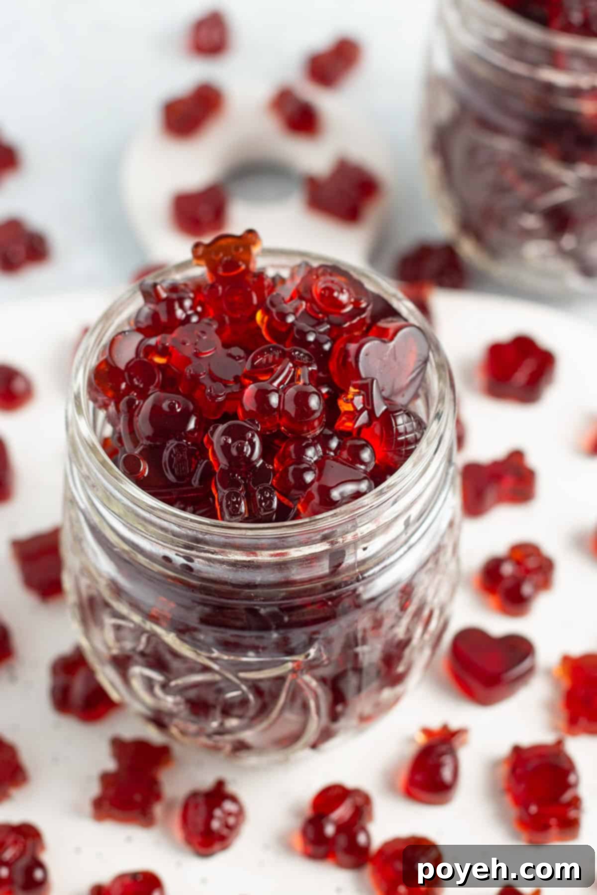 Sour Cherry Chews 3 Close-up, overhead, angled view of a glass jar overflowing with bright red tart cherry gummies on a white countertop.