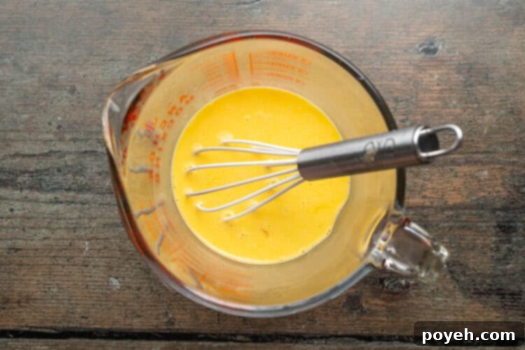 Overhead view of a glass measuring cup containing thoroughly whisked scrambled eggs and milk, with a silver-handled whisk resting inside, showing the pale and foamy texture.