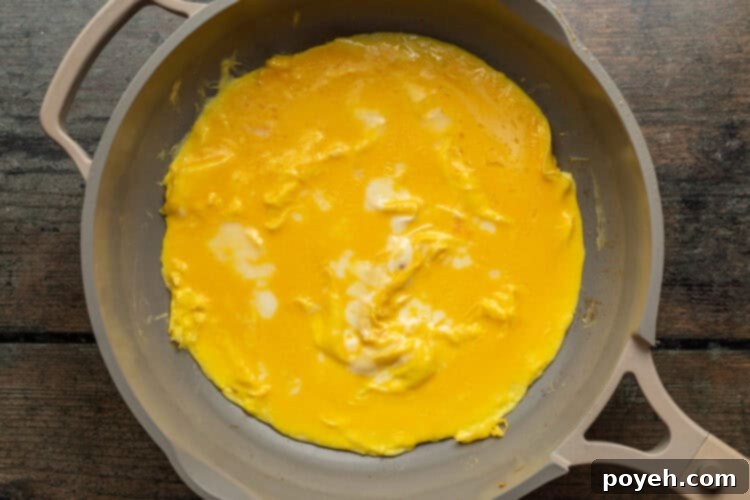 Overhead view of scrambled eggs carefully spread out across a hot skillet, forming the base of a Mexican omelette, with edges beginning to set.