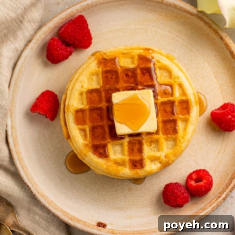 Overhead view of round frozen Eggo wafflse stacked on a plate and topped with maple syrup, butter, and raspberries.