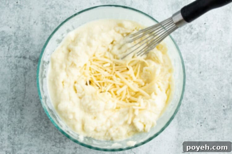 Overhead view of creamy potato mixture in a large bowl with shredded white cheese and a whisk.