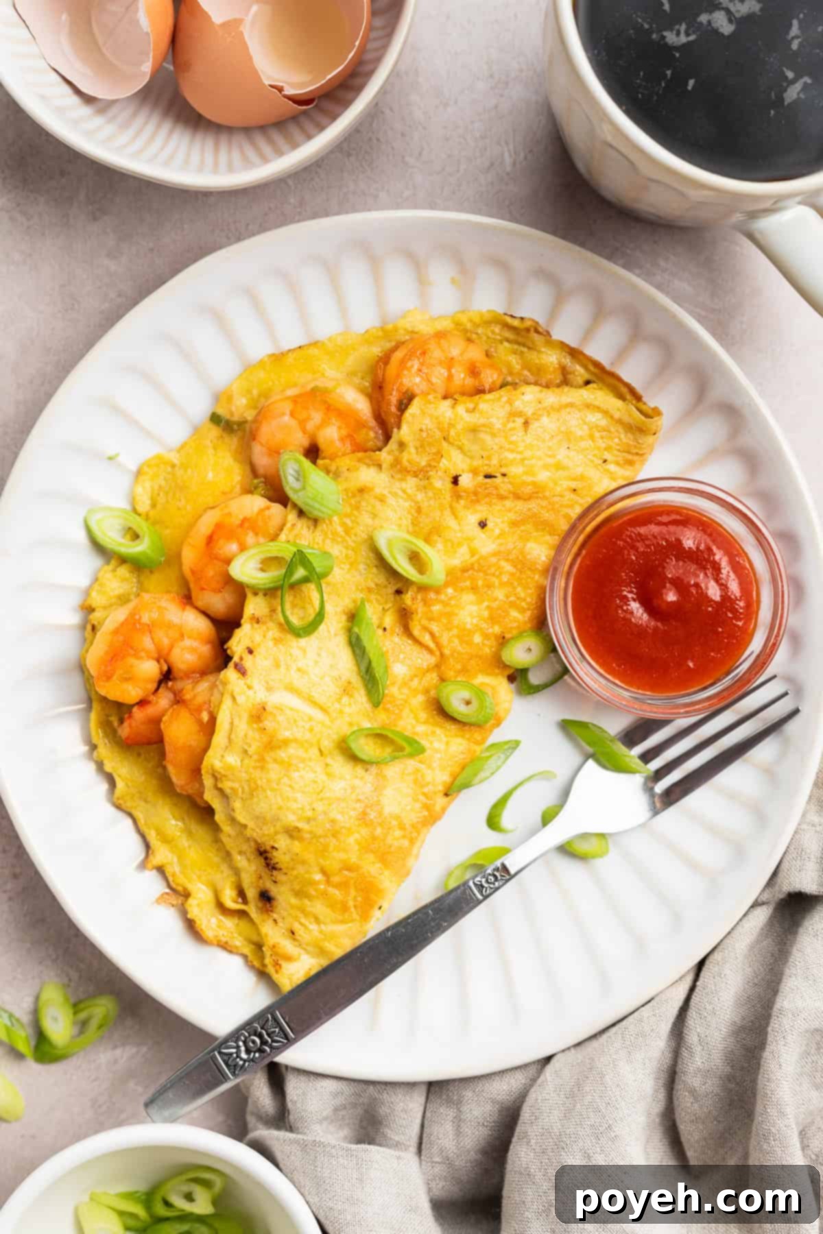 An overhead shot reveals a perfectly golden, half-moon shaped shrimp omelette, elegantly topped with a sprinkle of fresh chopped green onions, resting on a clean white plate. A tiny bowl of vibrant red ketchup offers a savory contrast beside it.