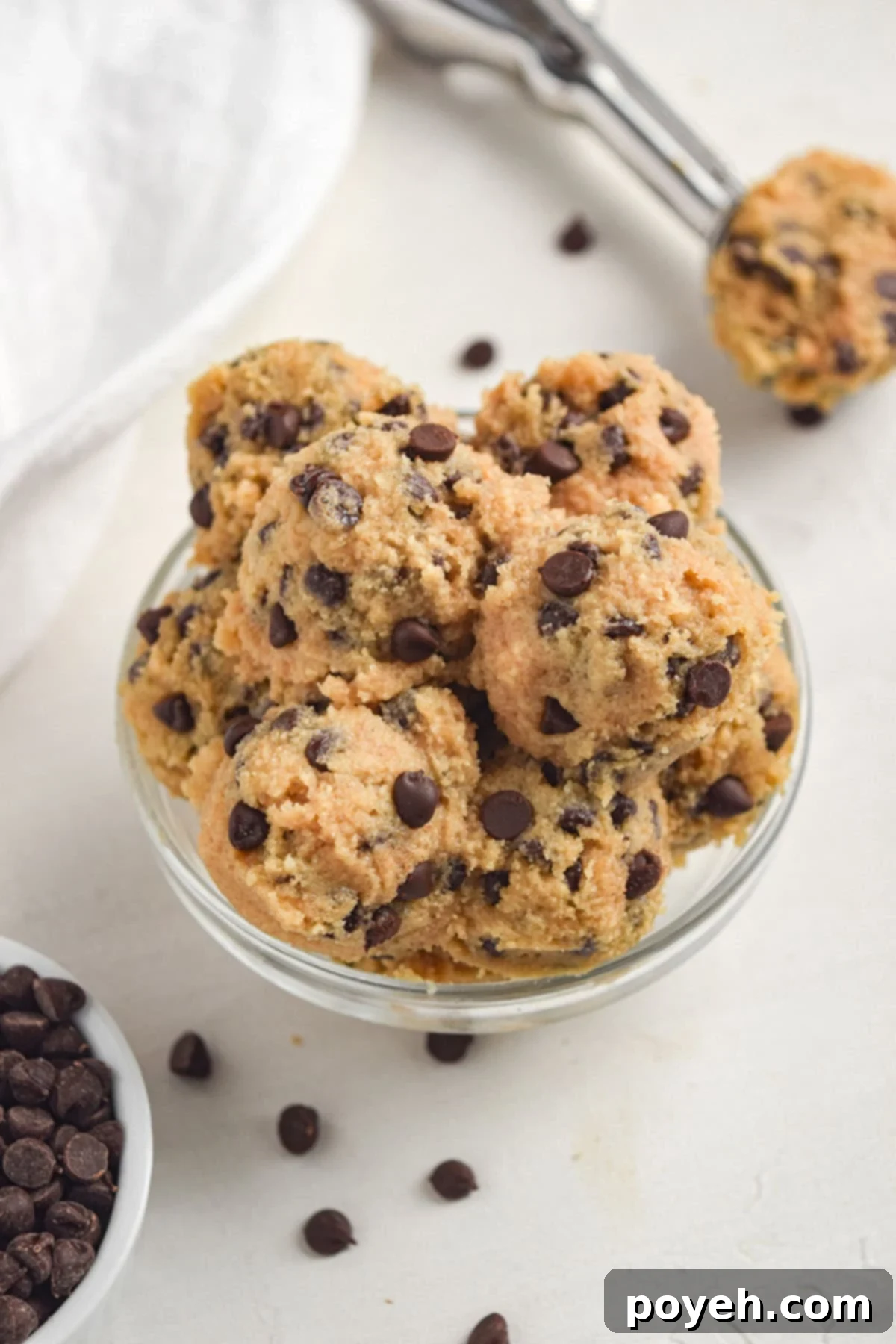 A glass mixing bowl holding scoops of keto chocolate chip cookie dough on a white tabletop with chocolate chips.