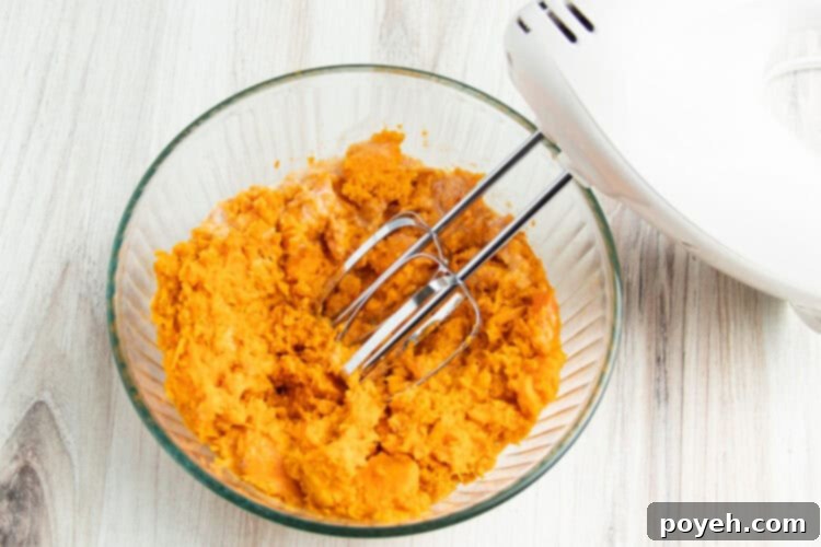 Overhead view of whipped sweet potatoes in a large glass mixing bowl with a white hand mixer fitted with beater attachments.