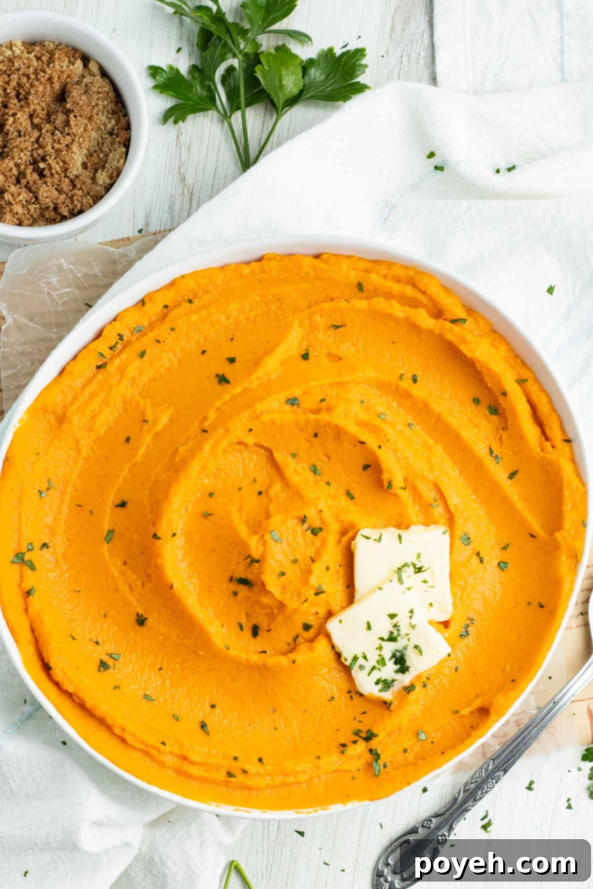 Overhead, zoomed-out view of a white bowl filled with bright orange whipped sweet potatoes on a white tablecloth. Potatoes are topped with 2 pats of butter and chopped chives.