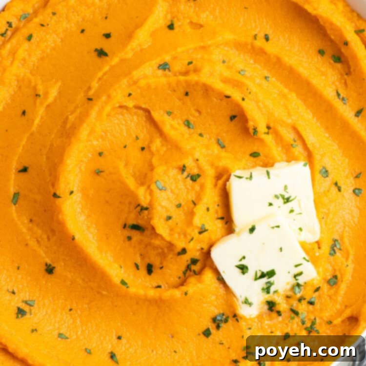 Overhead, zoomed-out view of a white bowl filled with bright orange whipped sweet potatoes on a white tablecloth. Potatoes are topped with 2 pats of butter and chopped chives.