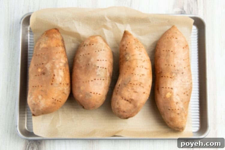 Overhead view of 4 large sweet potatoes lined horizontally on a baking sheet lined with parchment paper.