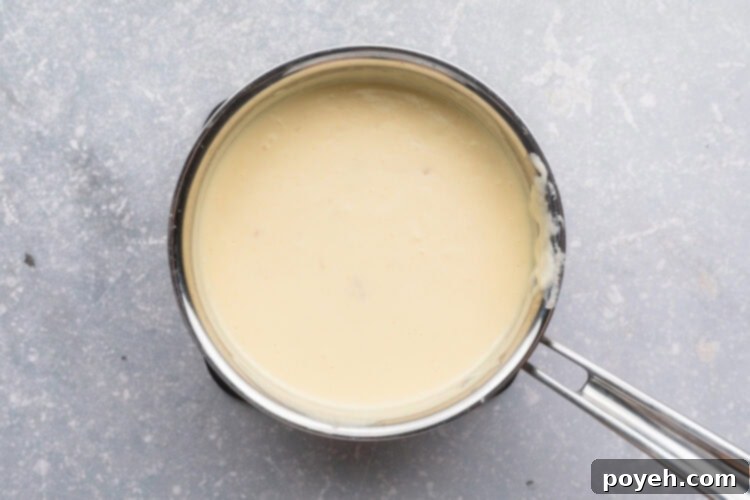 Overhead view of a small silver saucepan filled with a heavy cream sauce on a neutral blue-gray background.