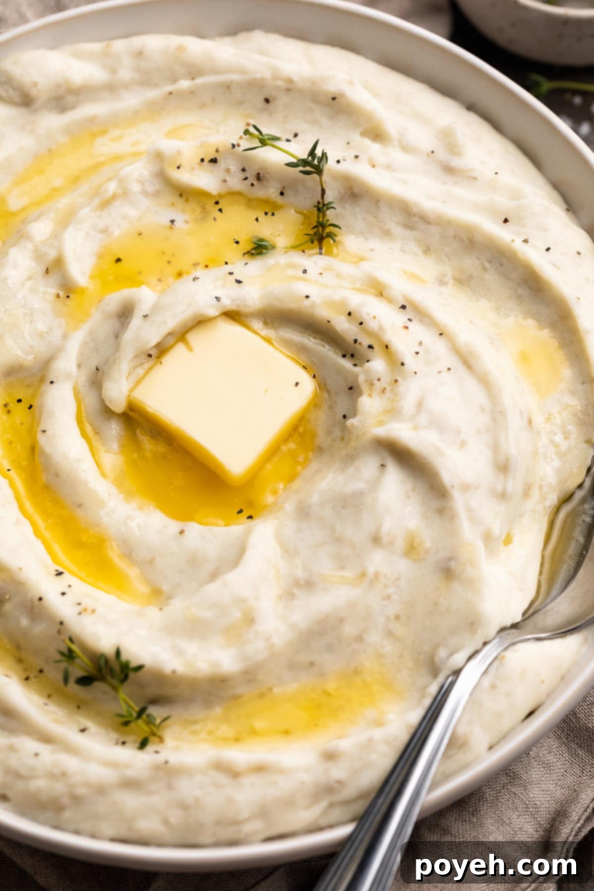 Velvety Whipped Potatoes 3 Close-up, slightly angled view of a large bowl holding perfectly whipped potatoes topped with a generous swirl of melted butter and a fresh pat of butter.