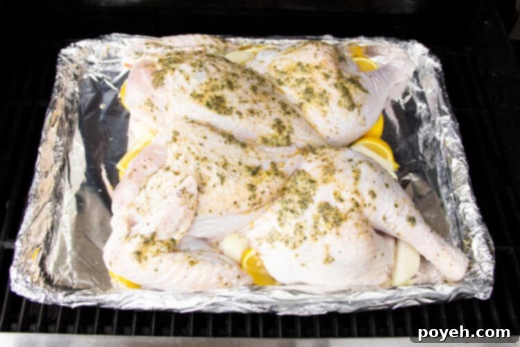 Angled, overhead view of a spatchcocked turkey covered in compound butter resting in an aluminum disposable pan lined with foil on a grill grate..