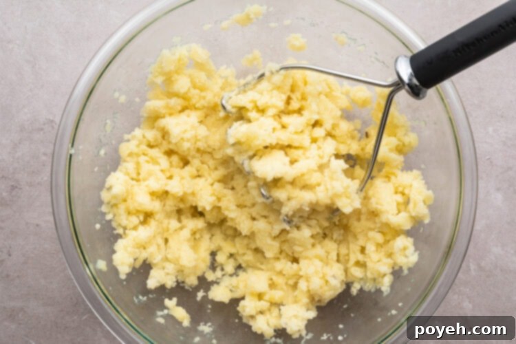 Overhead view of lightly mashed potatoes in a large glass mixing bowl on a neutral countertop, ready for additions.