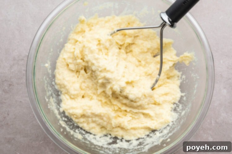 Overhead view of creamy mashed potatoes in a large glass mixing bowl with a potato masher resting on a neutral countertop, showing the finished texture.