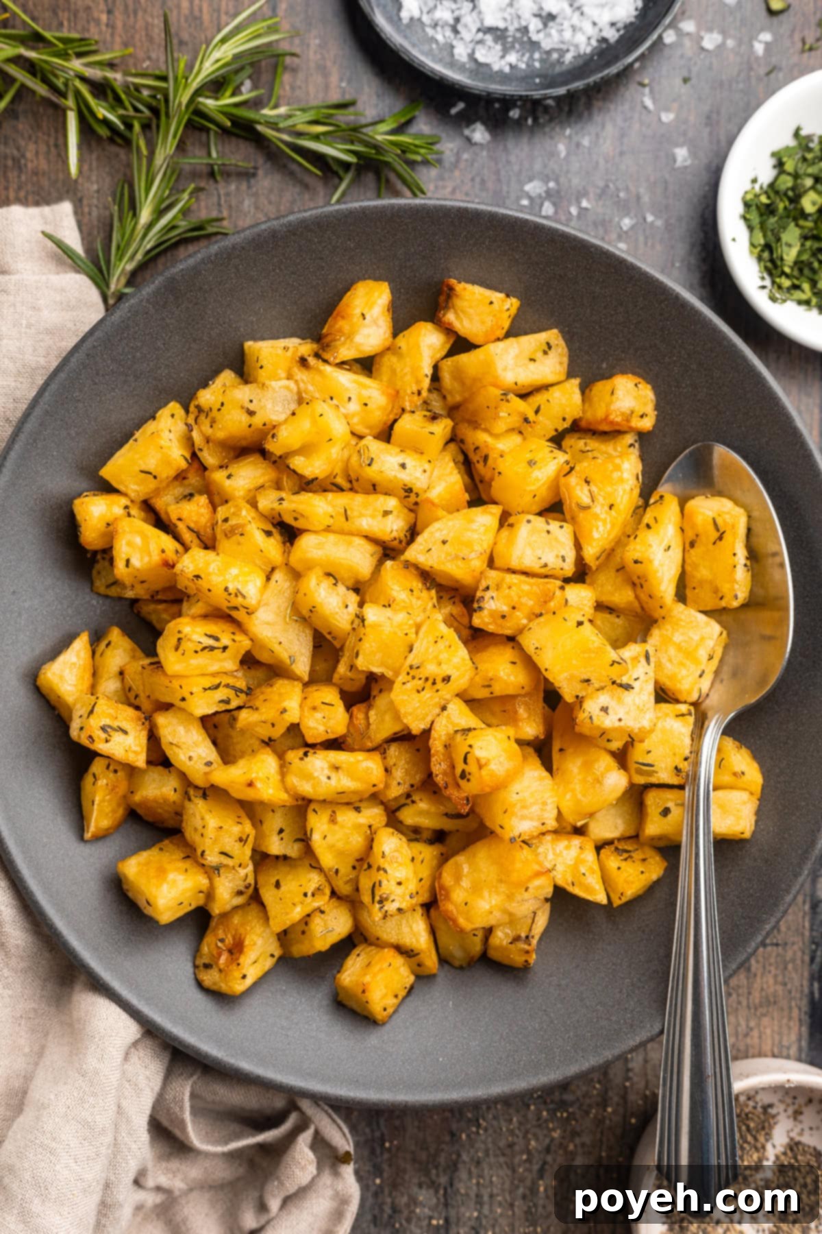 Overhead view of a clear glass bowl filled with diced potatoes, thoroughly tossed with dried herbs, neutral oil, salt, and pepper, glistening and ready for roasting.