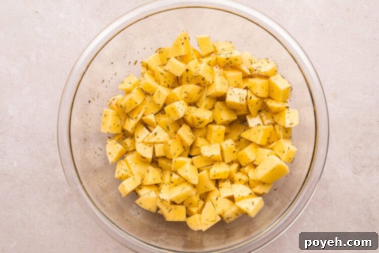 Overhead view of diced potatoes in a large glass mixing bowl, thoroughly coated with oil and seasonings, glistening and ready for the next step.