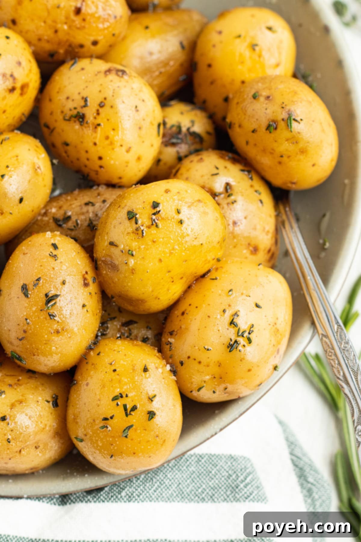 Overhead, close-up view of confit potatoes in a large bowl on a white tablecloth. Golden-brown potatoes are nestled amongst fresh rosemary and thyme sprigs.