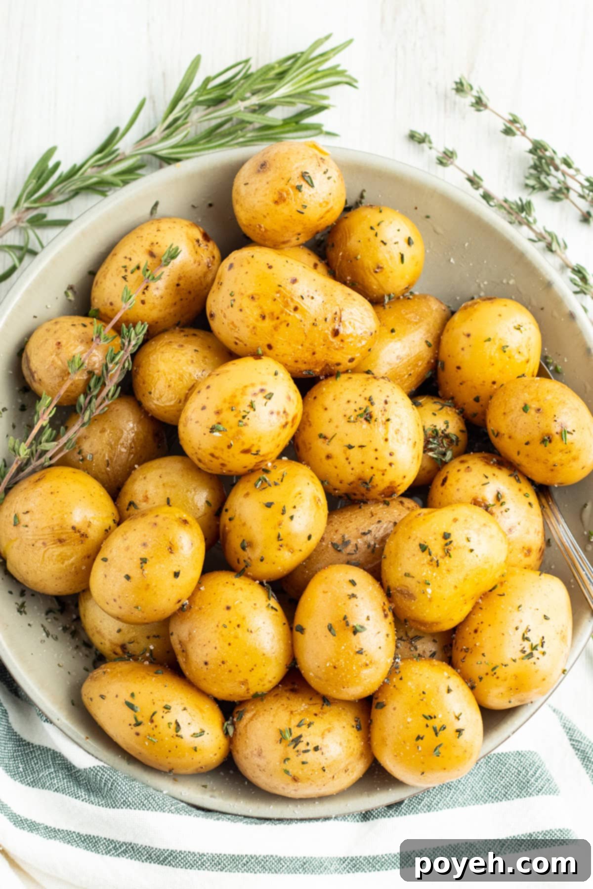 Overhead view of confit potatoes in a large bowl on a white tablecloth. The potatoes are glistening with oil and flecked with green herbs.