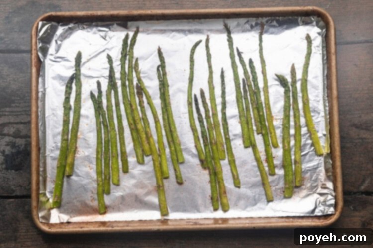 Asparagus spears evenly spread on a baking sheet lined with aluminum foil, seasoned and ready for roasting.