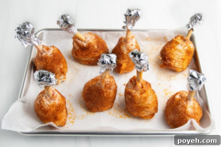 Lollipop chicken legs, with exposed bones covered by aluminum foil, standing upright on a baking sheet lined with parchment paper.