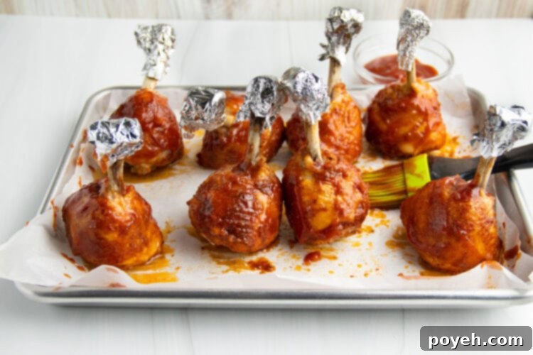Lollipop chicken legs, with exposed bones covered by aluminum foil, standing upright on a baking sheet lined with parchment paper. The chicken is generously coated in BBQ sauce.