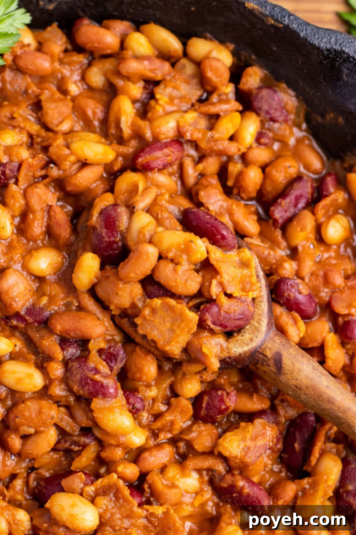 Close-up of smoked baked beans in a large cast-iron skillet with a large wooden spoon.