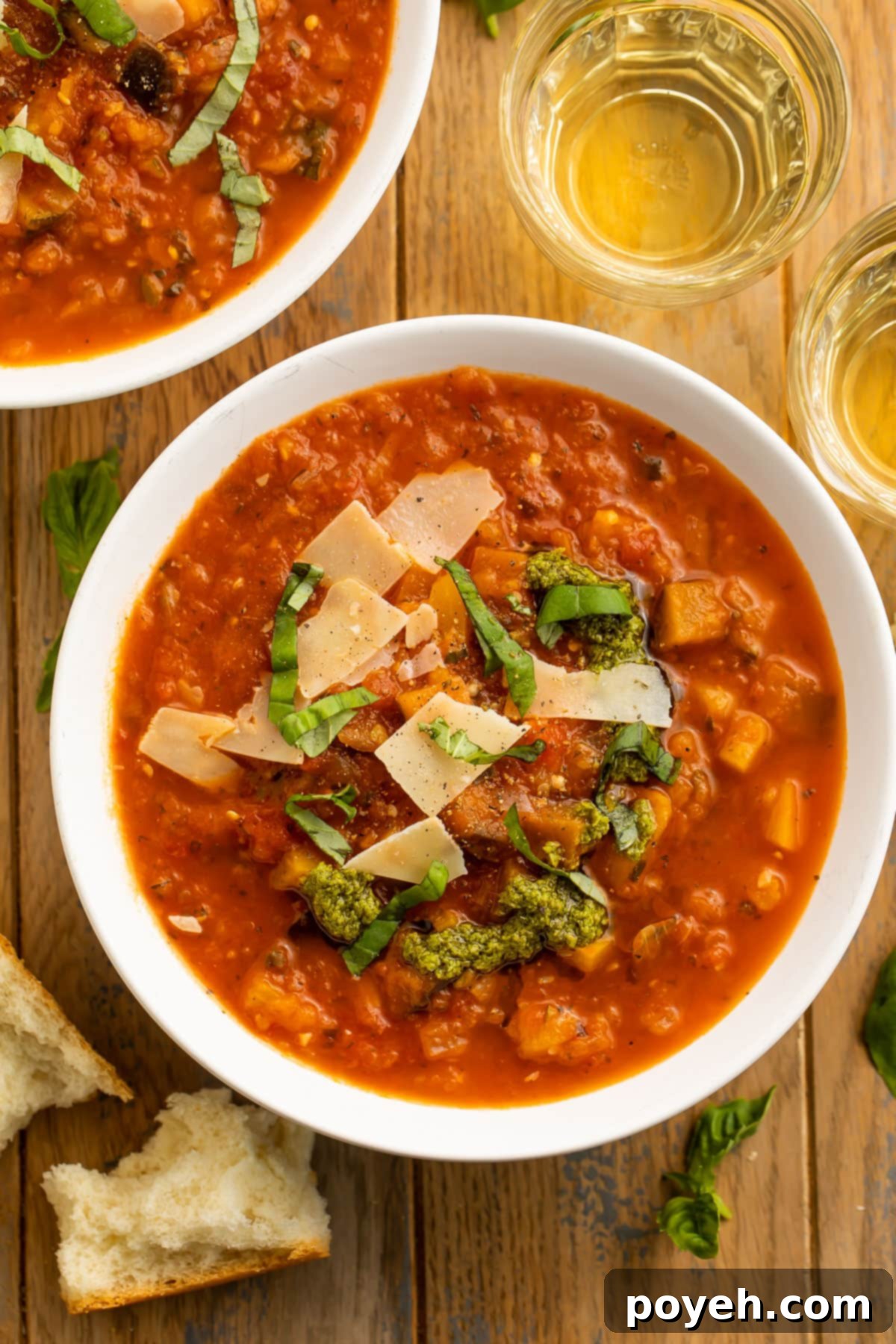 Top-down view of two bowls of ratatouille soup on a wooden table, garnished with fresh basil, alongside a glass of white wine and a French baguette.
