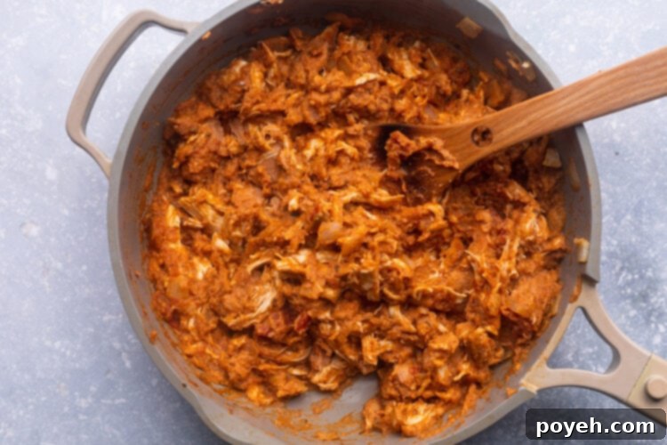 Shredded chicken, refried beans, salsa, taco seasoning, and onions in a large skillet with a wooden spoon.