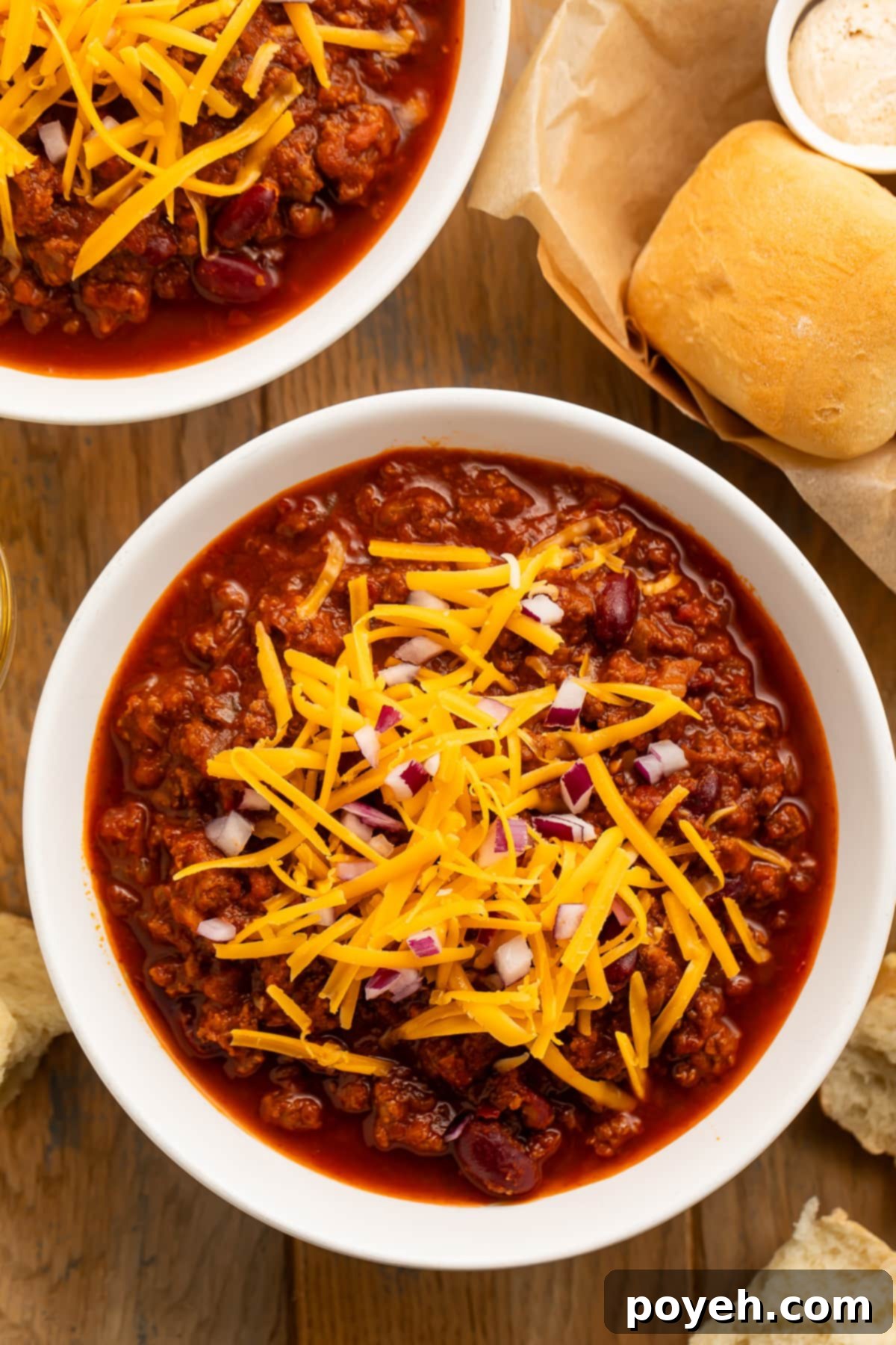 Overhead view of a bowl of dark red Texas Roadhouse chili topped with orange shreds of cheddar cheese and diced red onion on a dark tabletop.