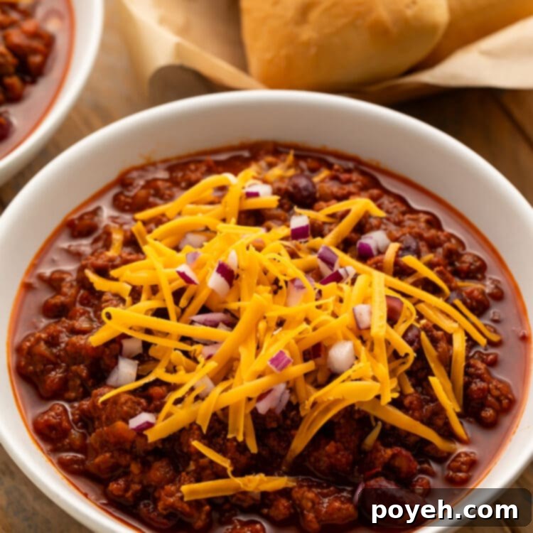 Angled view of a white bowl holding deep red chili topped with shredded cheddar cheese and diced red onions in front of a basket of rolls.