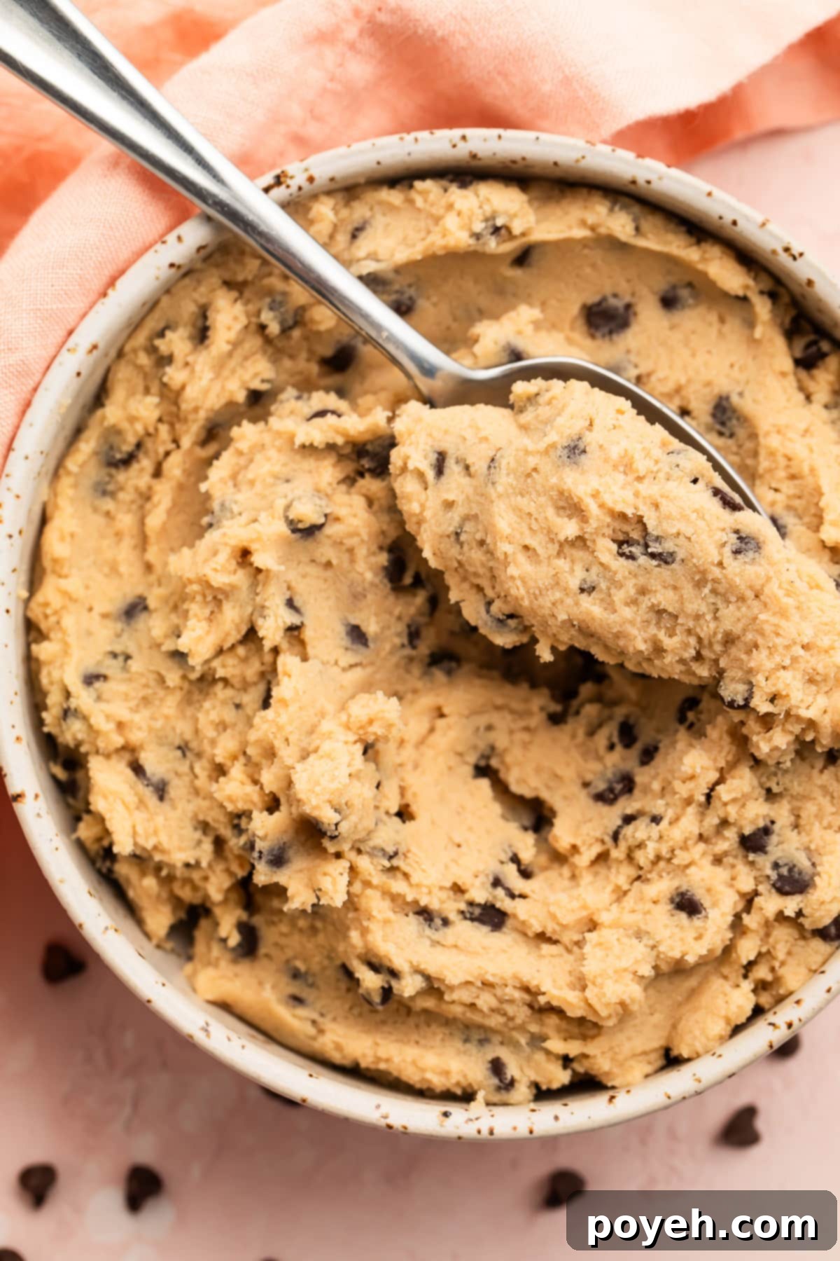 Gluten free cookie dough in a large white bowl with a spoon, surrounded by mini chocolate chips and a pink napkin.