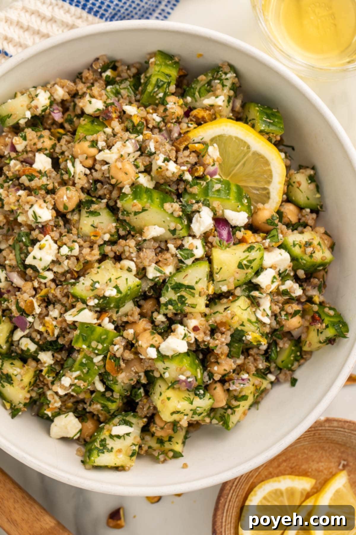 A large white bowl brimming with the viral Jennifer Aniston salad, featuring a colorful mix of quinoa, chickpeas, crisp cucumber, crunchy pistachios, creamy feta, and an abundance of fresh herbs. Slices of bright yellow lemon are casually placed around the bowl, adding a fresh visual appeal.