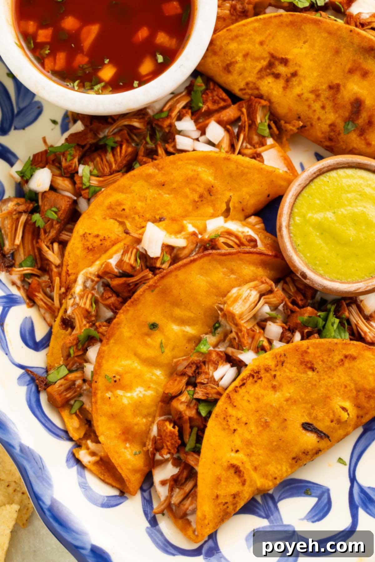 Four vegan birria tacos arranged on a blue and white platter with avocado sauce and salsa.