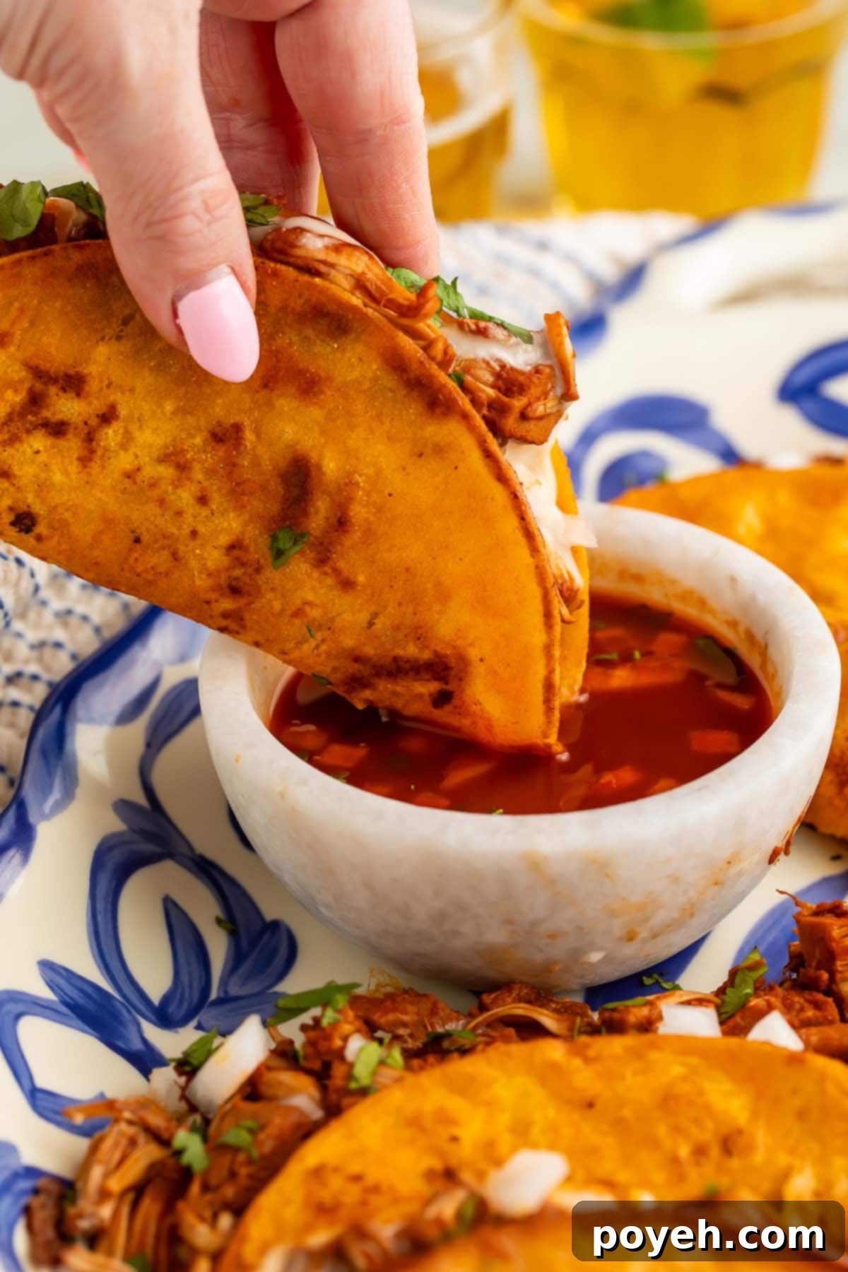 A vegan birria taco, held by a white woman's hand with light pink painted fingernails, as it's being dipped into a bowl of red salsa.