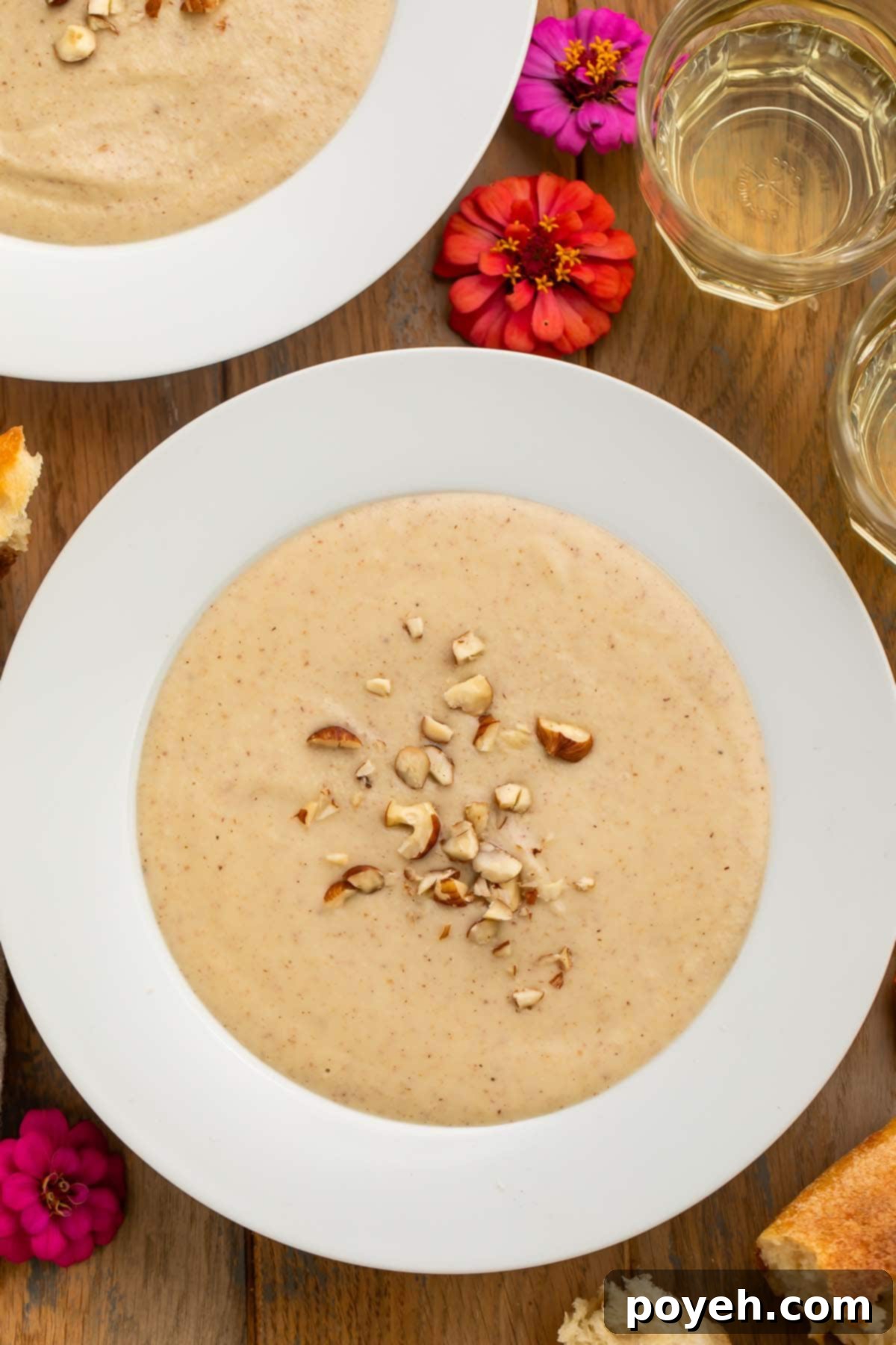 A large white bowl of creamy, pale hazelnut soup topped with chopped hazelnuts and fresh thyme, resting on a rustic wooden table adorned with delicate pink flowers, inviting warmth and comfort.