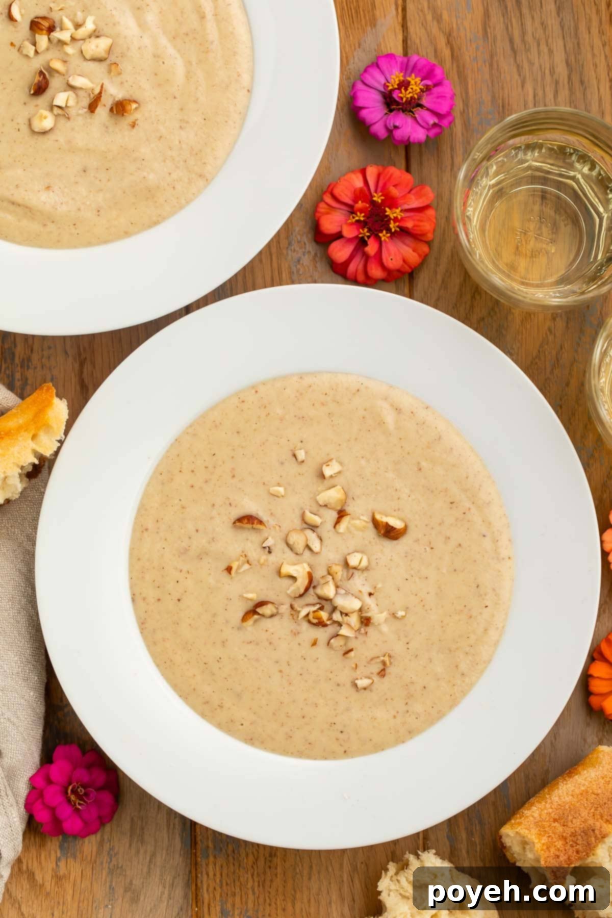 Close-up of creamy hazelnut soup, garnished with finely chopped hazelnuts and a sprig of fresh thyme, in a white bowl on a wooden surface with soft pink floral accents, highlighting its rich texture and inviting presentation.