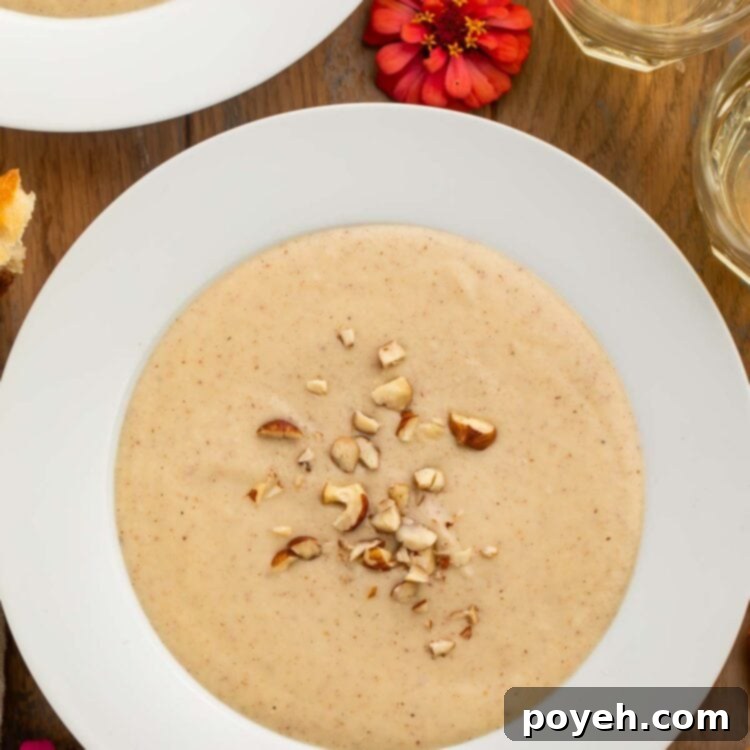 A large white bowl of creamy, pale hazelnut soup topped with chopped hazelnuts, on a wooden table with pink flowers.