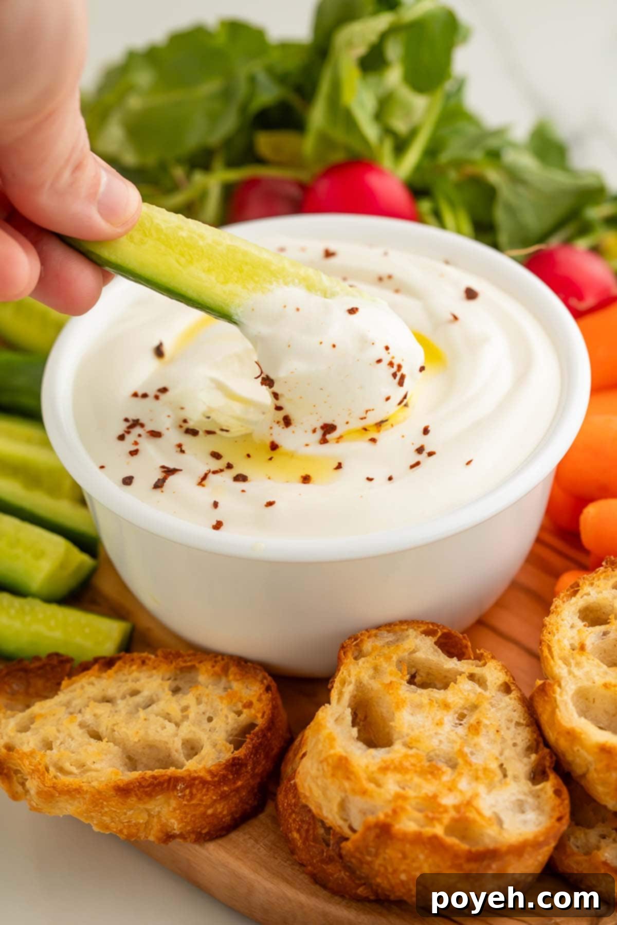 A white woman's hand holds a sliver of cucumber as she uses it to scoop whipped cottage cheese out of a bowl, showcasing its smooth and creamy texture.