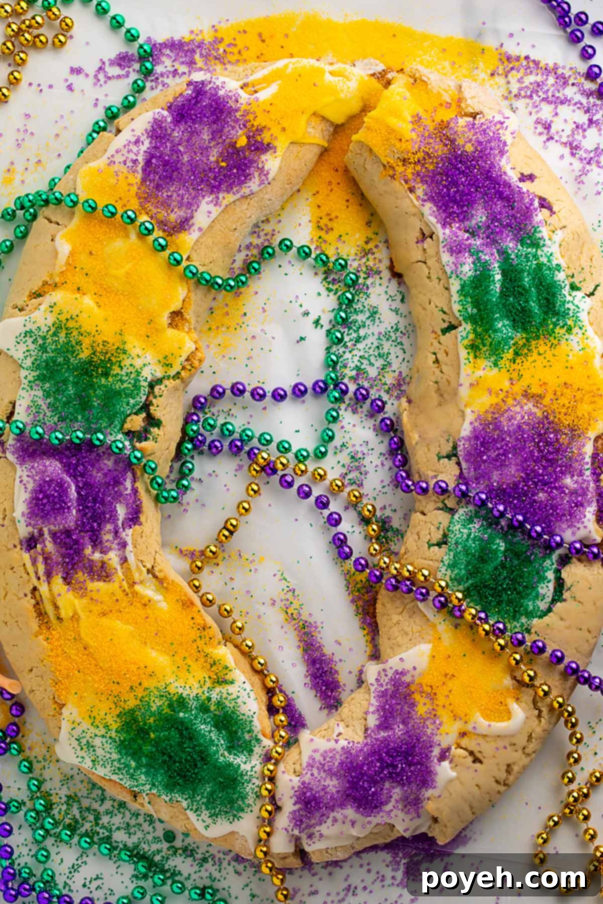 A large oval-shaped gluten free king cake topped with purple, yellow, and green sanding sugar and surrounding by purple, gold, and green Mardi Gras bead necklaces.